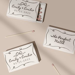 May include: Two white matchboxes with black calligraphy. One box says "Emily & Emilio 05.25.2025" with a black bow. The other box says "The Perfect Match".