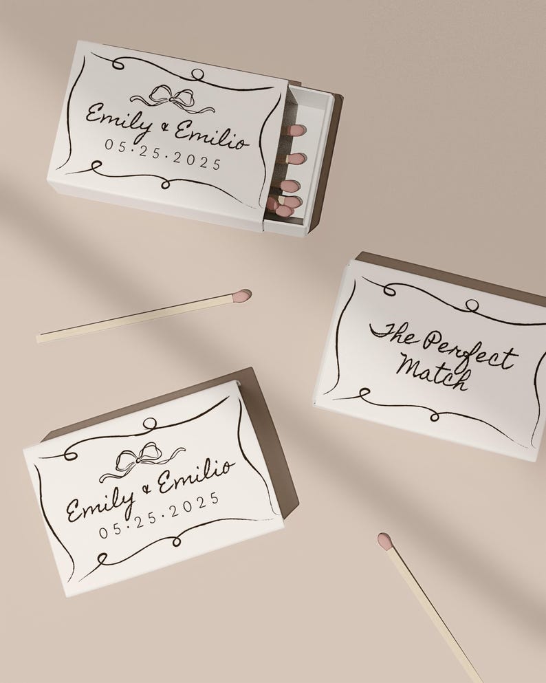 May include: Two white matchboxes with black calligraphy. One box says "Emily & Emilio 05.25.2025" with a black bow. The other box says "The Perfect Match".