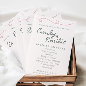 Puede incluir: Tarjetas de programa de ceremonia de boda con un diseño de espiral rosa. Las tarjetas están apiladas en una caja de madera y presentan los nombres "Emily & Emilio" y el orden de la ceremonia. La fecha es el 25 de mayo de 2025, en Southland.