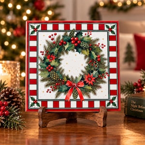 May include: A square ceramic tile featuring a central Christmas wreath illustration with green pine needles, red berries, pinecones, and poinsettias, tied with a red ribbon. The border has a red and white striped pattern with holly accents.
