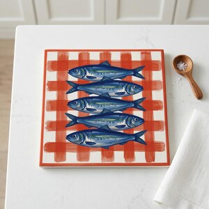 May include: Square ceramic serving tray featuring a red and white checkered pattern. The tray is decorated with five blue fish illustrations. A wooden spoon with salt and a white napkin are next to the tray.