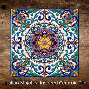 May include: Square ceramic tile with a colorful Italian Majolica design. The tile features a central floral medallion in shades of blue, orange, and yellow, surrounded by intricate scrollwork and floral motifs. The text "Italian Majolica Inspired Ceramic Tile" is at the bottom.