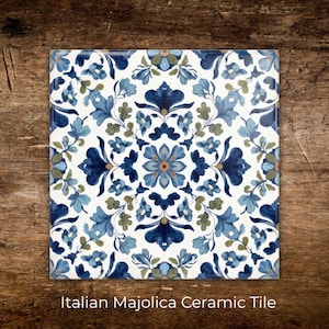 May include: Square ceramic tile with a floral design in shades of blue, green, and white. The tile features a symmetrical pattern of stylized flowers and leaves. The text "Italian Majolica Ceramic Tile" is at the bottom.