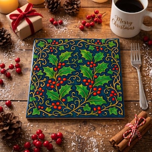 May include: A square ceramic tile featuring a dark blue background with a repeating pattern of green holly leaves, red berries, and gold swirling vines. The tile is displayed on a wooden surface alongside a gift box, pinecones, and a mug.