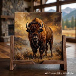 May include: A ceramic tile featuring a detailed painting of a bison in a natural landscape. The tile is displayed in a wooden stand. The bison is brown with black horns, set against a backdrop of mountains and a cloudy sky. The tile measures 6x6 inches.