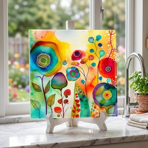 May include: A colorful floral art print on a white easel. The artwork features vibrant blooms in shades of blue, red, yellow, and green. The painting is displayed on a white easel, set against a window background.
