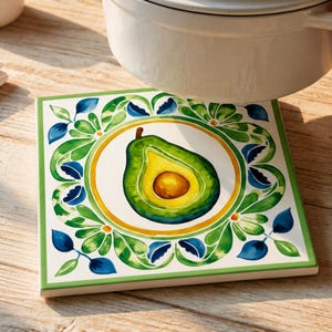 May include: Square ceramic trivet with a watercolor avocado design. The avocado is green and yellow, centered on a white background, surrounded by a yellow ring and green and blue floral accents. A white pot rests on the trivet.