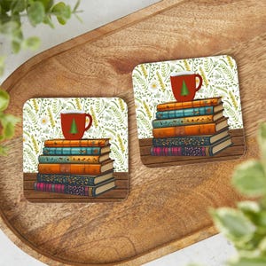 May include: Two square coasters with a stack of books and a red mug with a Christmas tree design. The coasters have a floral background with a wooden base. The books are in various colors, including orange, blue, and burgundy.