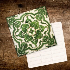 May include: Two square ceramic tiles. One tile features a green and white floral pattern with gold accents. The other tile is white with a brick pattern. The tiles are displayed on a wooden surface.