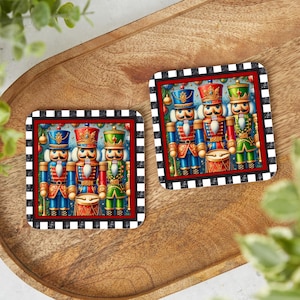 May include: Two square coasters with a black and white checkered border and a red inner border. Each coaster features a colorful illustration of three toy nutcracker soldiers in blue, red, and green uniforms.