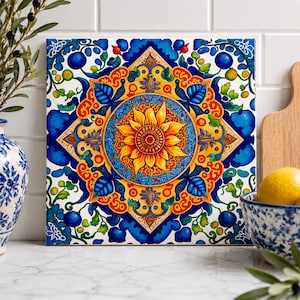 May include: A square ceramic tile featuring a vibrant central yellow sunflower motif surrounded by intricate blue, orange, and green floral and geometric patterns. The design includes symmetrical swirling vines, leaves, and small berries set against a white background.