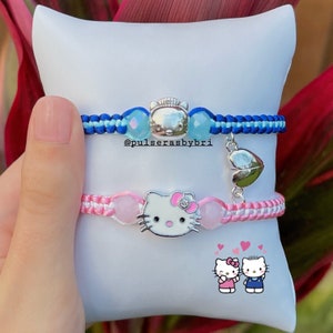 May include: Two friendship bracelets with Hello Kitty charms. The blue bracelet has a light blue bead and a silver charm. The pink bracelet has a pink bead and a silver charm. The bracelets are on a white background with a small image of two Hello Kitty characters.