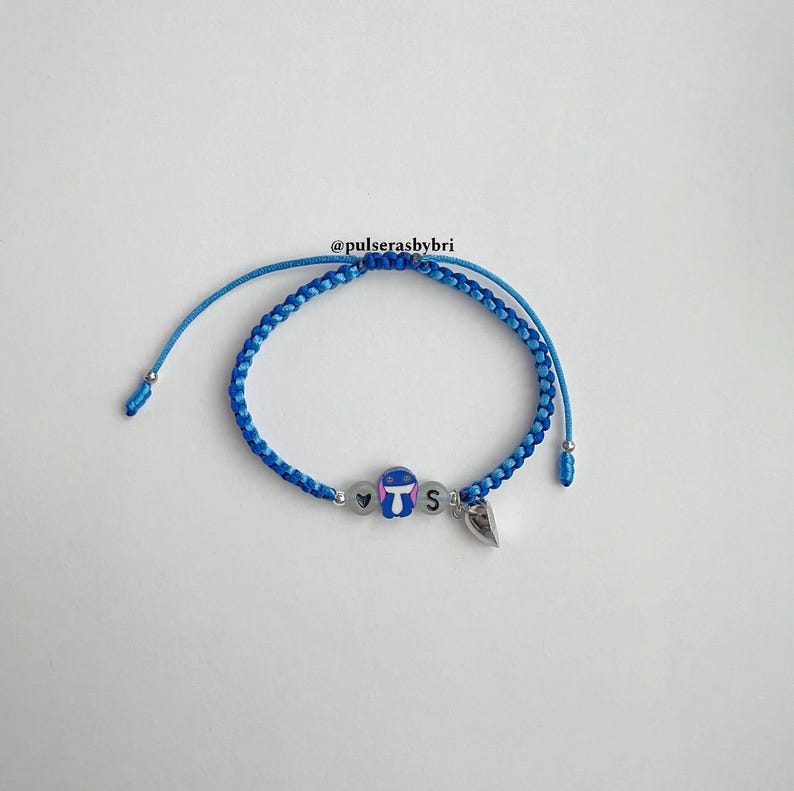Pode incluir: Pulseira de amizade tran&ccedil;ada azul e azul claro com um pequeno amuleto Stitch, um amuleto de cora&ccedil;&atilde;o e um amuleto da letra 'S'.