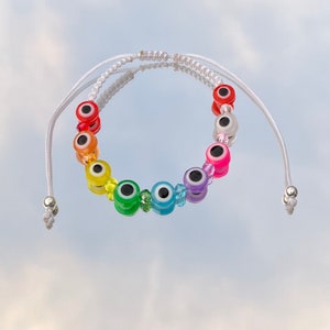 May include: A white string bracelet with rainbow colored evil eye beads. The beads are arranged in a rainbow pattern, with red, orange, yellow, green, blue, indigo, and violet beads.