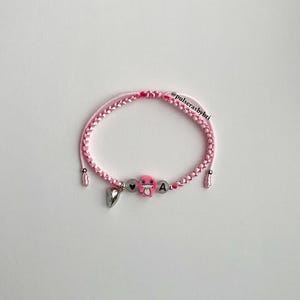 Pode incluir: Pulseira de corda tran&ccedil;ada rosa e branca com um amuleto de sapo rosa, um amuleto de cora&ccedil;&atilde;o prateado e um amuleto de letra A prateada. A pulseira tem o texto "@pulserasbybri" nela.