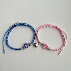 Pode incluir: Duas pulseiras de amizade com cord&otilde;es azuis e rosa, um fecho de cora&ccedil;&atilde;o prateado e contas de letras que soletram "T.S" e "A".