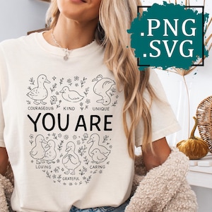 May include: Ivory-colored t-shirt with a black graphic of ducks and the words "YOU ARE." The design includes words like "courageous," "kind," and "loving." The shirt is a casual, everyday wear item.