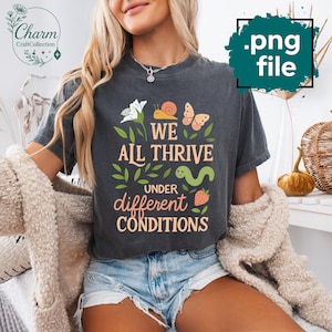 May include: Dark gray t-shirt with the text "WE ALL THRIVE UNDER DIFFERENT CONDITIONS" in a whimsical design featuring a snail, butterfly, worm, strawberry, and flowers. The shirt has a vintage, worn look.