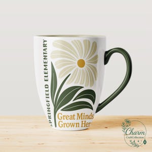 May include: White ceramic mug with a green handle and rim. The mug features a floral design with a daisy and leaves in shades of beige and green. Text on the mug reads "Springfield Elementary" and "Great Minds Grown Here."
