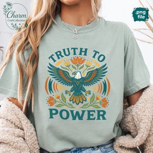 May include: A light green t-shirt with the text "TRUTH TO POWER" and a graphic of an eagle with outstretched wings. The eagle is surrounded by floral elements in orange, green, and teal. The shirt has short sleeves.