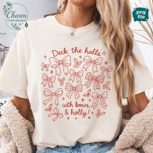 May include: A cream-coloured t-shirt featuring a red festive design. The graphic reads "Deck the halls with bows & holly!" and includes illustrations of bows and holly. The t-shirt is a relaxed fit, ideal for the Christmas season, and is made of a soft material.