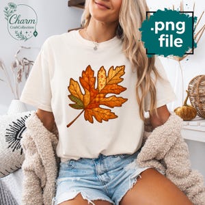 Könnte beinhalten: Cremefarbenes T-Shirt mit einem großen, farbenfrohen Blattdesign. Das Blatt ist ein Patchwork aus orangefarbenen, gelben und grünen Mustern mit einem karierten Mittelteil. Das Shirt hat den Text "Charm CraftCollection" in einem kreisförmigen Logo.