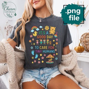 Puede incluir: Camiseta gris oscuro con el texto "IT'S A GOOD DAY TO CARE FOR TINY HUMANS" en letras y gráficos coloridos. El diseño incluye un arcoíris, un pato, un oso de peluche, flores y niños. La camiseta es un archivo .png.