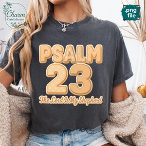 May include: Dark gray t-shirt with the text "PSALM 23" in a light brown, textured font. The number "23" is in a similar font with heart patterns. The phrase "The Lord Is My Shepherd" is below the numbers.