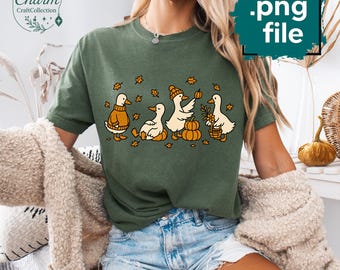 May include: Green t-shirt featuring a whimsical design of four ducks in autumn attire. The ducks are depicted with pumpkins, leaves, and a basket of flowers. The shirt has a relaxed fit and short sleeves.