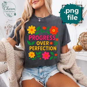Puede incluir: Camiseta gris oscuro con el texto "PROGRESS OVER PERFECTION" en rosa y amarillo, rodeado de flores coloridas y hojas verdes. La camiseta tiene un aspecto vintage, ligeramente descolorido.