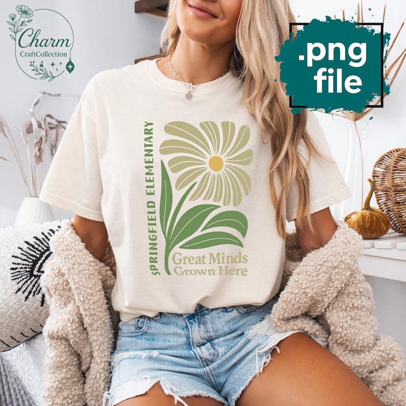 May include: Cream-colored t-shirt featuring a large floral design in shades of green and yellow. The text "Springfield Elementary" is printed vertically, with "Great Minds Grown Here" below the flower. The shirt has short sleeves.