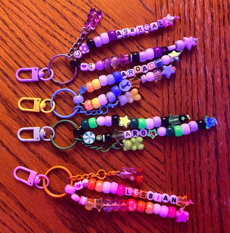 May include: A selection of colourful beaded keychains. Each keychain spells out a different word using letter beads, such as "ASEXUAL," "AROACE," and "LESBIAN." The keychains are adorned with star and heart charms and are attached to metal clasps.