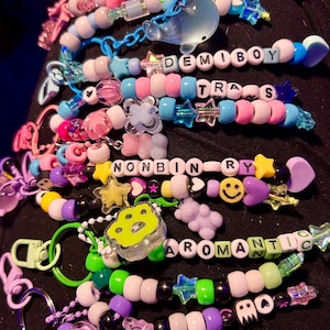 May include: A selection of colourful beaded keychains, each spelling out words like "DEMIGIRL", "DEMIBOY", "TRANS", "NONBINARY", "AROMANTIC", and "ACE". The keychains incorporate diverse beads, charms, and star-shaped embellishments.