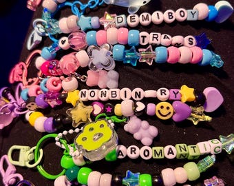 May include: A collection of beaded keychains featuring various pastel and vibrant plastic beads, star-shaped charms, and bear-shaped pendants. The beads spell out terms including DEMIGIRL, DEMIBOY, TRANS, NONBINARY, AROMANTIC, and ACE, with metal clips.
