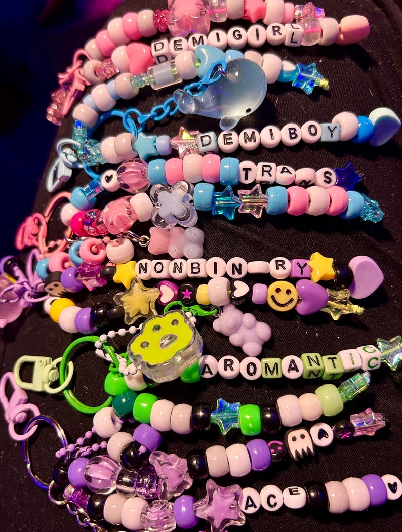 May include: A selection of colourful beaded keychains, each spelling out words like "DEMIGIRL", "DEMIBOY", "TRANS", "NONBINARY", "AROMANTIC", and "ACE". The keychains incorporate diverse beads, charms, and star-shaped embellishments.