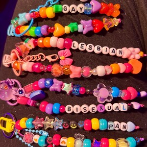 May include: A selection of vibrant beaded bracelets. Each bracelet spells out a word: "GAY", "LESBIAN", "BISEXUAL", and "PAN" using white letter beads. The bracelets are adorned with various charms and shapes.