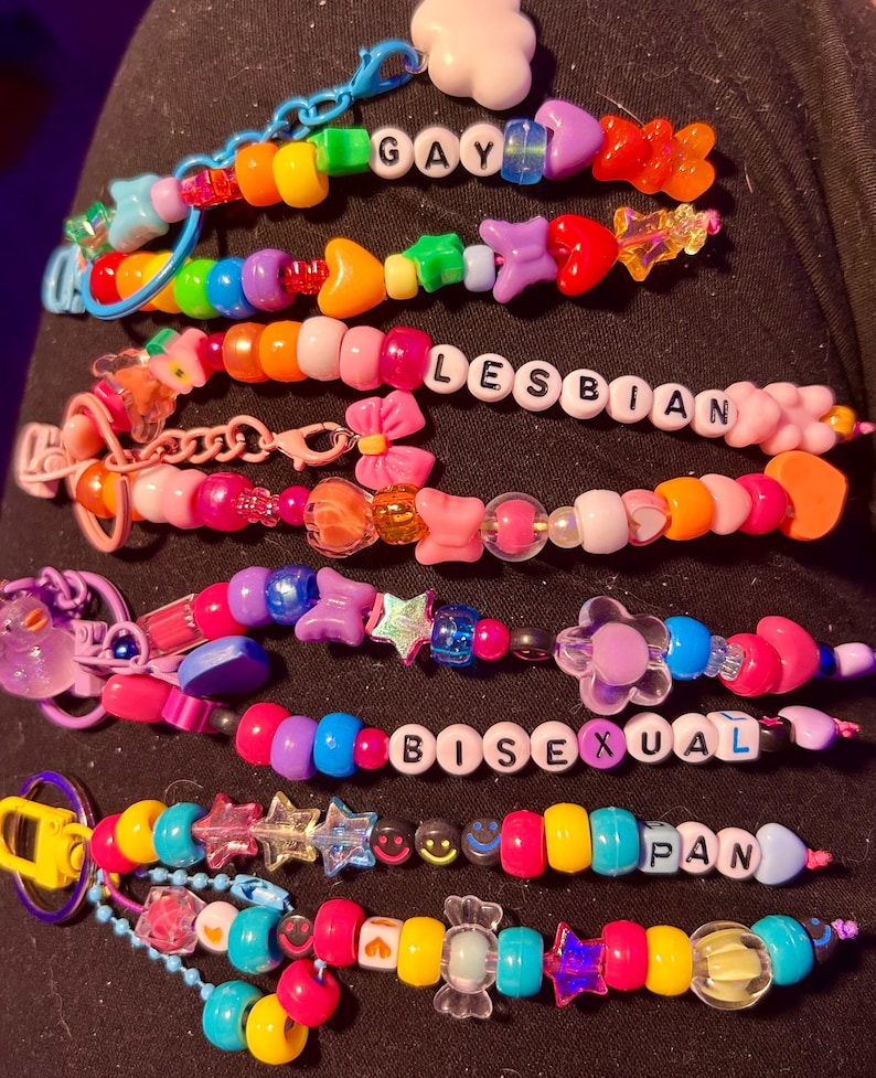 May include: A selection of vibrant beaded bracelets. Each bracelet spells out a word: "GAY", "LESBIAN", "BISEXUAL", and "PAN" using white letter beads. The bracelets are adorned with various charms and shapes.