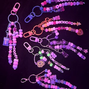 May include: A selection of vibrant beaded keyrings, each spelling out words like "Bisexual," "Lesbian," "Ace," "Aromantic," and "Trans." The keyrings incorporate diverse shapes and colours, such as stars, hearts, and bear charms.