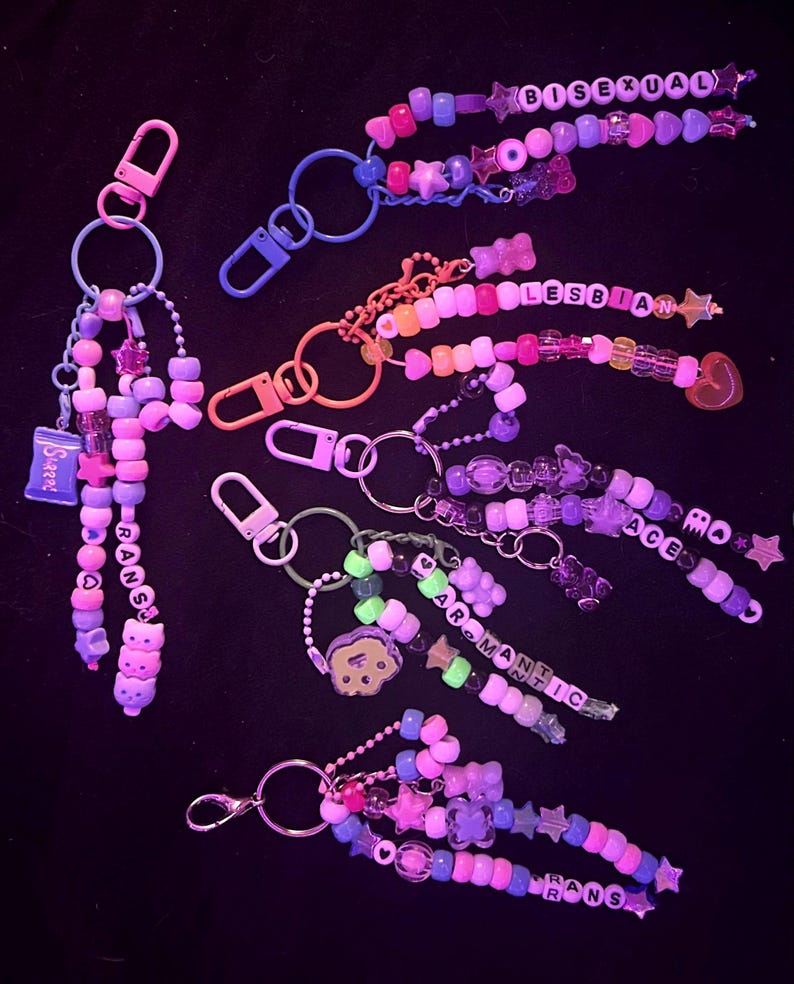 May include: A selection of vibrant beaded keyrings, each spelling out words like "Bisexual," "Lesbian," "Ace," "Aromantic," and "Trans." The keyrings incorporate diverse shapes and colours, such as stars, hearts, and bear charms.