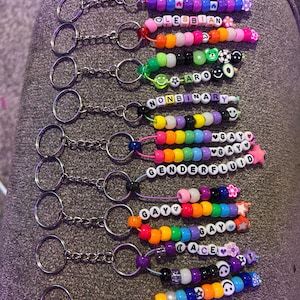 May include: A selection of beaded keyrings, each spelling out a word related to LGBTQ+ pride using colourful letter beads. Keyrings include the words: bisexual, lesbian, aromantic, nonbinary, genderfluid, gay, ace.