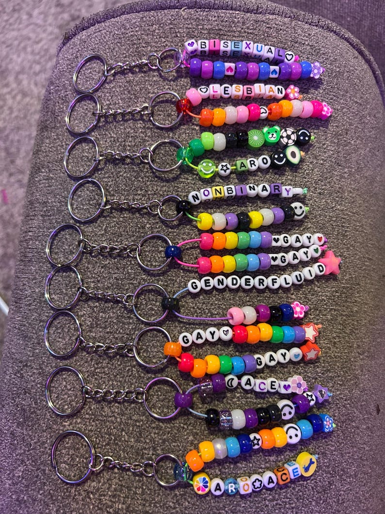 May include: A selection of beaded keyrings, each spelling out a word related to LGBTQ+ pride using colourful letter beads. Keyrings include the words: bisexual, lesbian, aromantic, nonbinary, genderfluid, gay, ace.