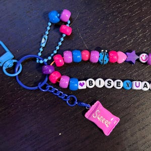 May include: A blue keyring with colourful beads spelling "BISEXUAL". The beaded design includes a ladybug, stars, and smiley faces. A small pink charm in the shape of a sweet wrapper is attached.