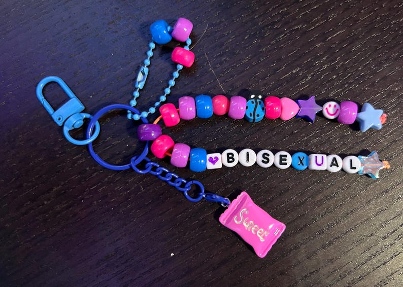 May include: A blue keyring with colourful beads spelling "BISEXUAL". The beaded design includes a ladybug, stars, and smiley faces. A small pink charm in the shape of a sweet wrapper is attached.