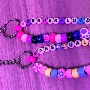 May include: Three beaded keychains with the words "GENDERFLUID" and "PANSEXUAL" spelled out in letter beads. The keychains feature a variety of colourful beads, including pink, purple, blue, and yellow, with decorative charms.