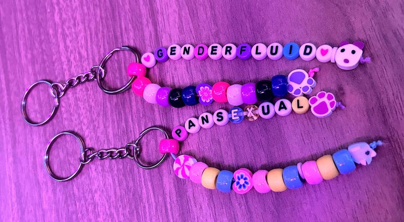 May include: Three beaded keychains with the words "GENDERFLUID" and "PANSEXUAL" spelled out in letter beads. The keychains feature a variety of colourful beads, including pink, purple, blue, and yellow, with decorative charms.