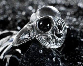 May include: A silver ring with a detailed skull design and a black gemstone eye. The ring is set against a black background.