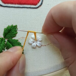 Puede incluir: Primer plano de una mano cosiendo una flor blanca con un tallo amarillo a un trozo de tela. La flor est&aacute; rodeada de hojas verdes y una fresa roja.