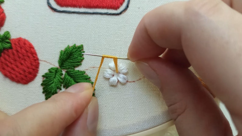 Puede incluir: Primer plano de una mano cosiendo una flor blanca con un tallo amarillo a un trozo de tela. La flor est&aacute; rodeada de hojas verdes y una fresa roja.