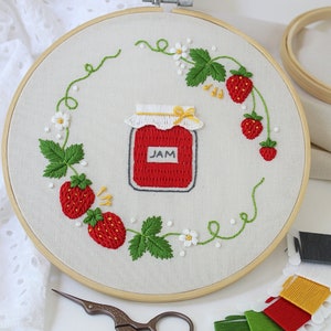 Puede incluir: Arte de bordado en aro con un tarro de mermelada de fresa roja con tapa blanca y la palabra "JAM" en la etiqueta. El tarro est&aacute; rodeado de una corona de hojas verdes, fresas rojas y flores blancas.