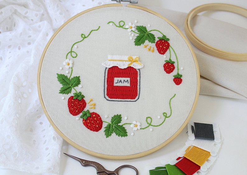 Puede incluir: Arte de bordado en aro con un tarro de mermelada de fresa roja con tapa blanca y la palabra "JAM" en la etiqueta. El tarro est&aacute; rodeado de una corona de hojas verdes, fresas rojas y flores blancas.